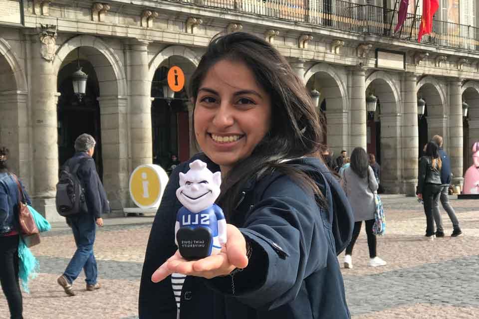Female Madrid student holding hand out with squishy Billiken, with arches in background